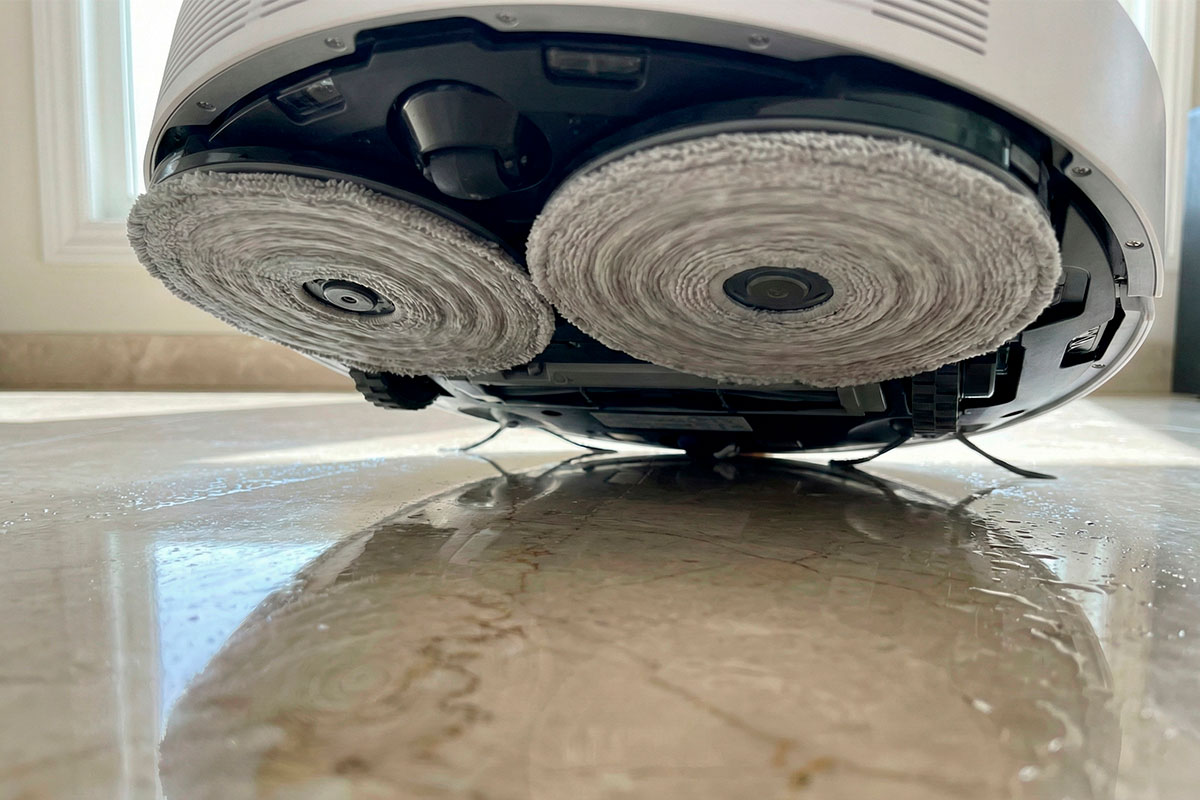 Primer plano de las mopas giratorias húmedas de una aspiradora trapeadora fregando un piso de mármol con reflejo.