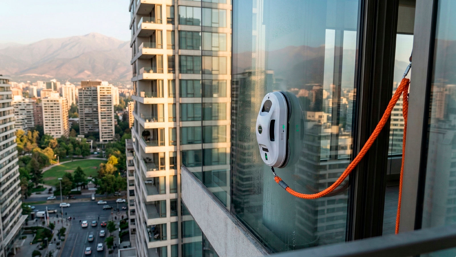 Robot limpiavidrios automático de Nuvac adherido a un ventanal de gran altura con cable de seguridad de color naranja brillante visible, con vista panorámica de la ciudad de Santiago de Chile de fondo.
